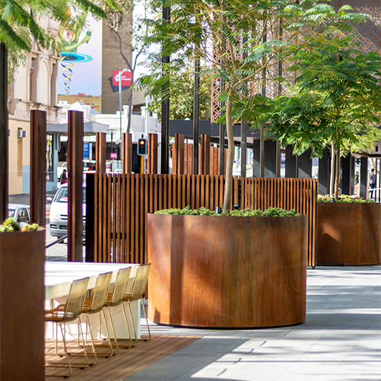 Grande jardinière circulaire en acier Corten accueillant un arbre dans un aménagement urbain contemporain.