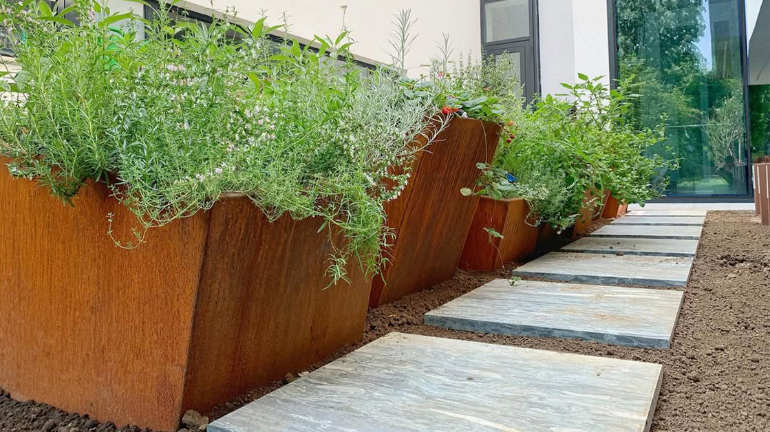 Alignement de jardinières en acier Corten sur mesure, garnies de plantes aromatiques le long d’une allée paysagère contemporaine.
