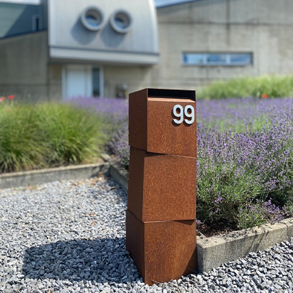 Borne d’adresse en acier Corten avec numéro métallique, installée dans un aménagement paysager contemporain.