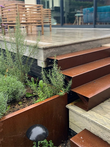 Escalier extérieur et bordure paysagère en acier Corten intégrés à une terrasse en bois entourée de végétation.