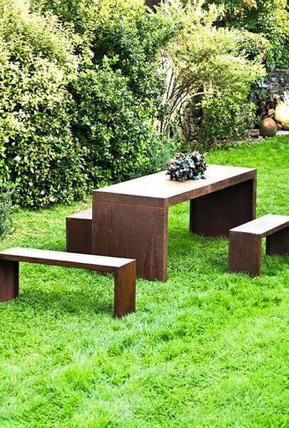 Ensemble composé d’une table rectangulaire et de bancs en acier Corten installé dans un jardin verdoyant.